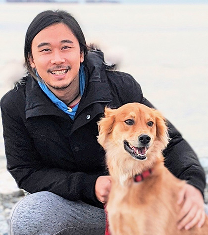 A man poses, crouched down beside his sitting dog in the snow.