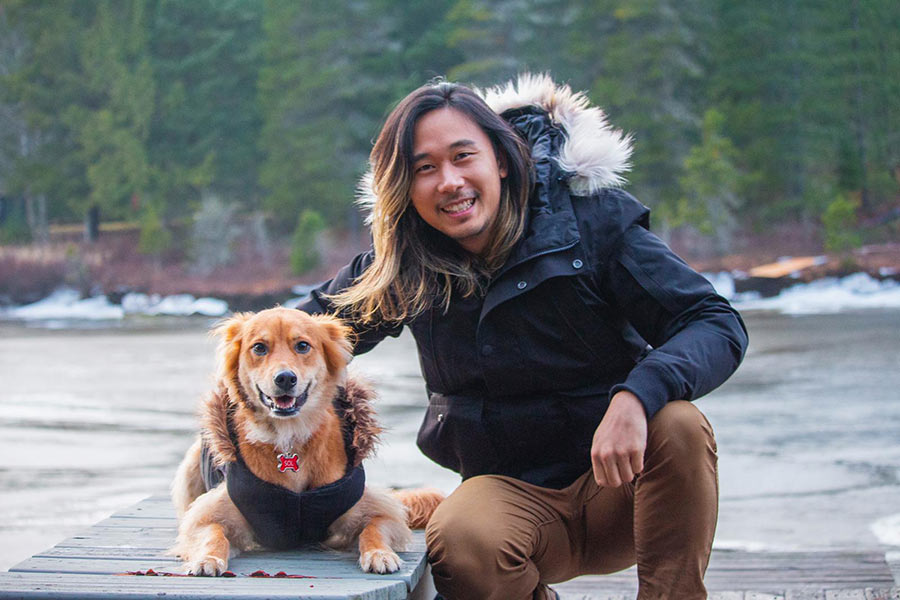 A man poses, crouched down beside his lying dog by a lake.